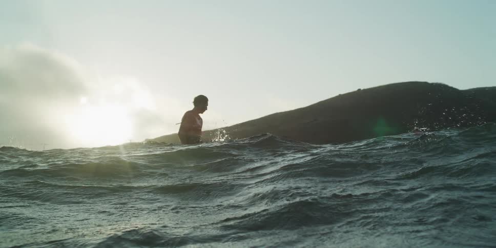 Man in the middle of the sea surfing a small wave with sun reflection on the horizon