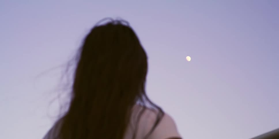 A person with his back and long hair is watching the moon in the sky on a day