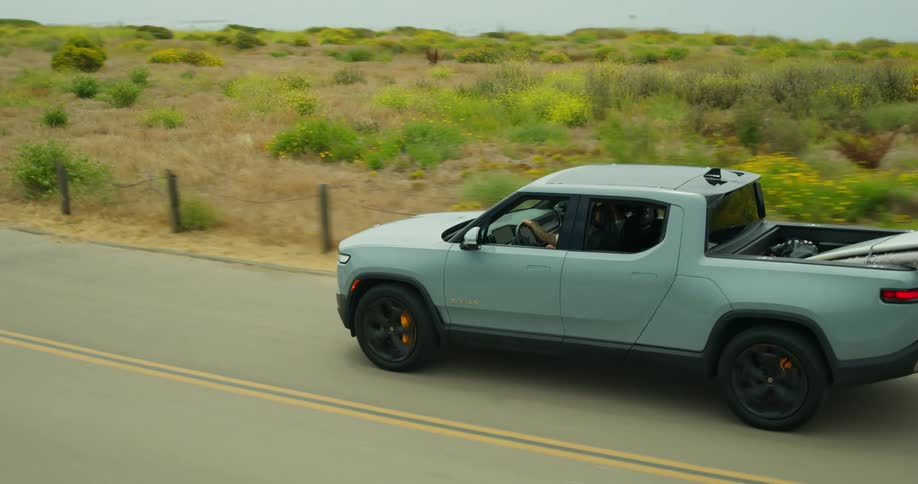 A pickup truck driving on an asphalt street with a large board on the back and the sea behind