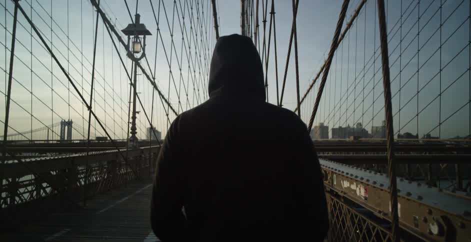 A man in a hoodie is walking on a pedestrian bridge at dusk