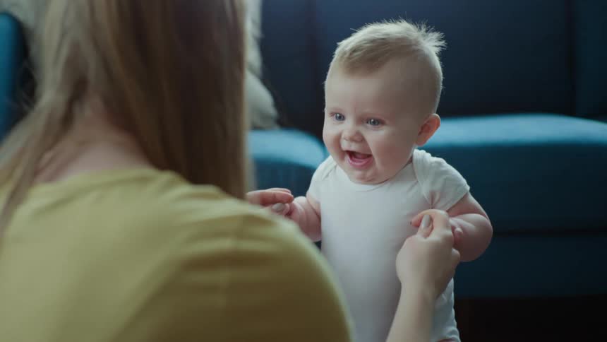 Mother with brown hair and a yellow t shirt holds toddlers hands near the blue couch in the living room