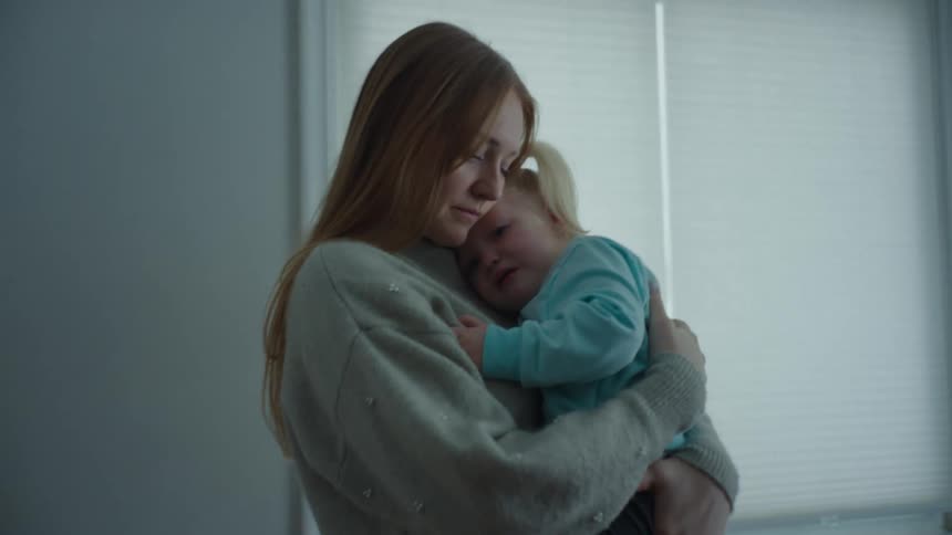 Mother with brown hair and a gray sweater holds toddler girl in the room