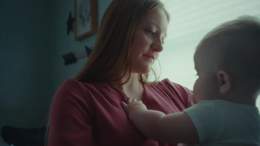 Mother with brown hair and red blouse holds her baby in her arms in a room