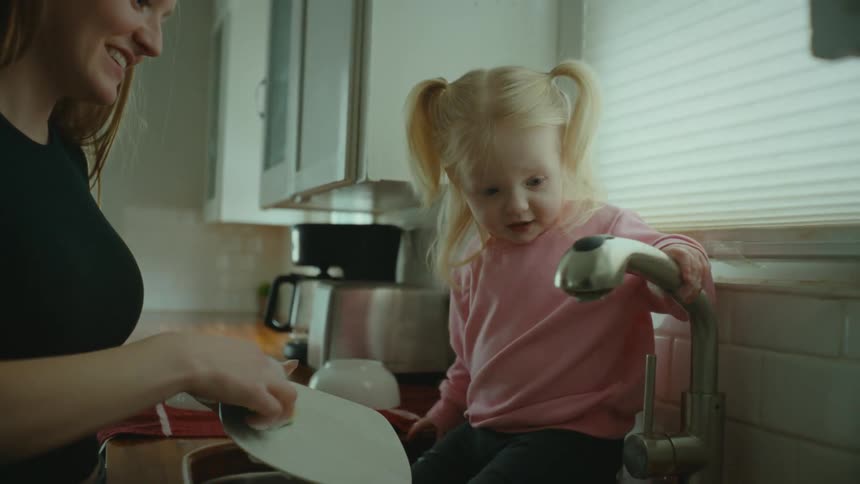 A little blonde girl in a pink shirt is in the sink helping her mother wash the dishes