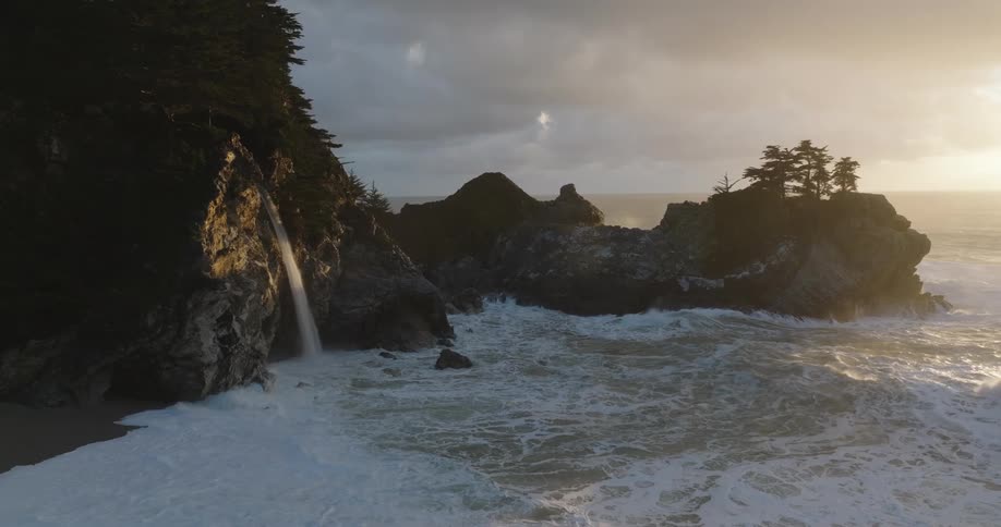 Aerial of a waterfall on a coastal mountain on a beach during sunset