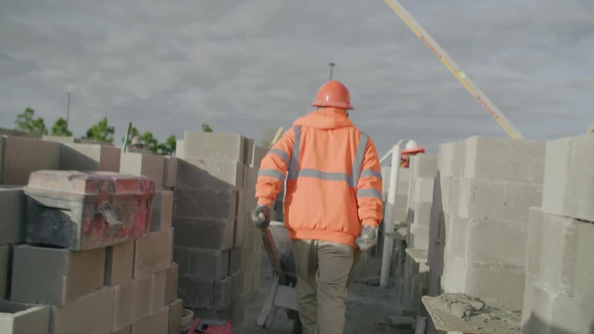 A worker in orange clothes and helmet pushing a cart in the middle of a construction site full of bricks