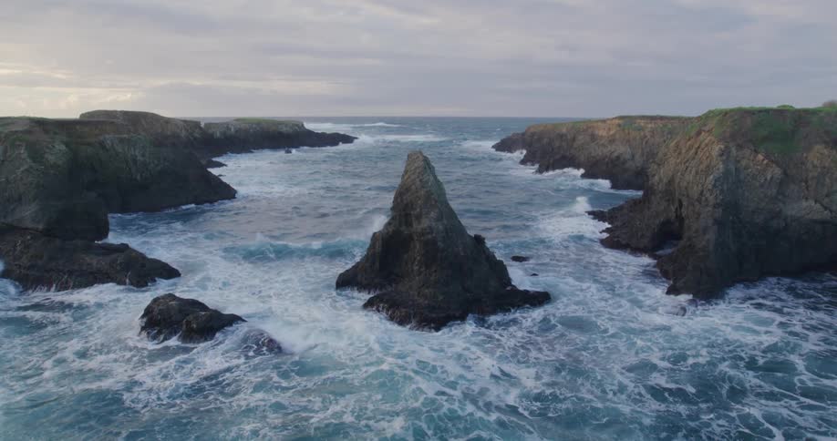 Waves crashing into a jagged rock in an ocean cove on a cloudy morning