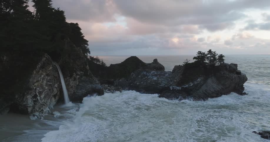 Waves crash into coastal rocks with a waterfall under a cloudy skyat sunset