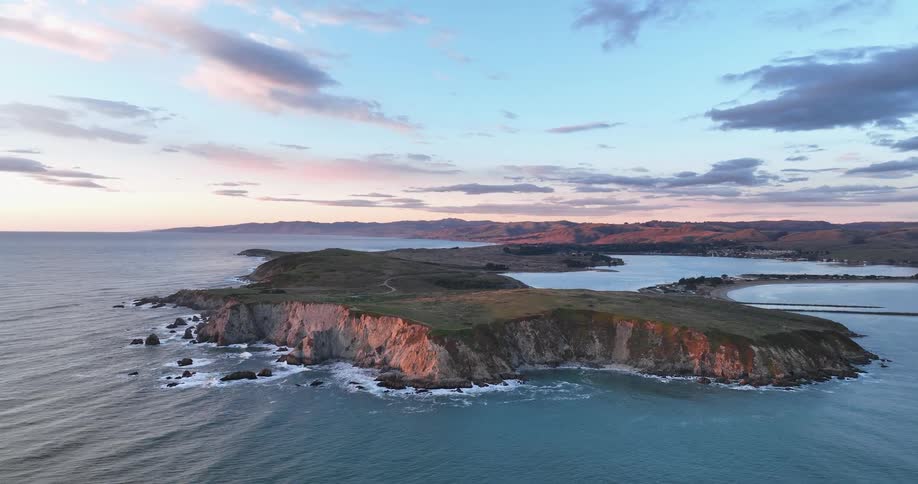 Aerial panoramic of an island peninsula surrounded by ocean water under a blue sky with scattered clouds and pink in the distance at sunset