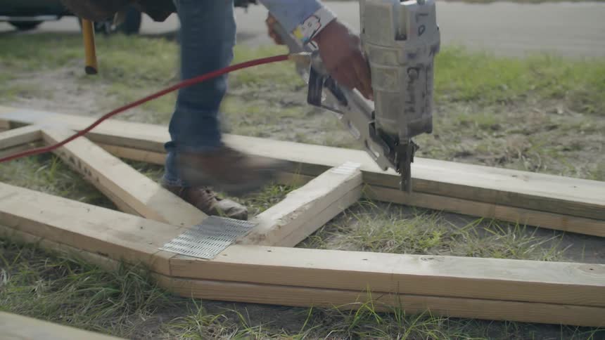 Construction worker pounds nails with an air gun into a wood shed frame laid flat on a lawn