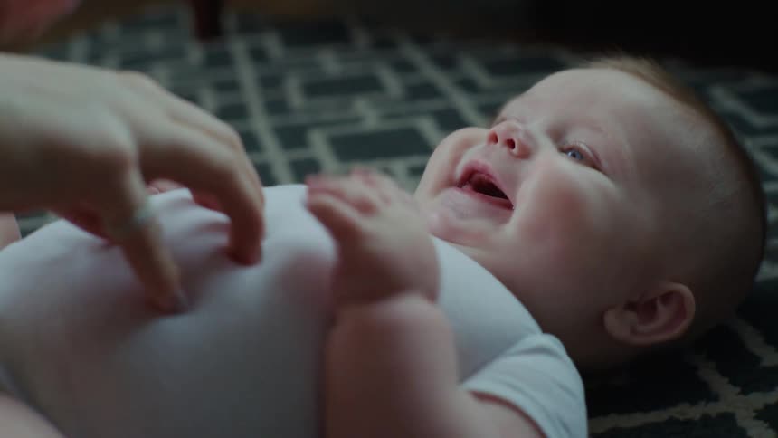 A little baby in a white shirt and blue eyes is lying down and laughing while someone tickles his belly
