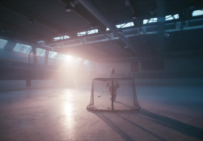 Hockey player holding a stick and pushing a goal across an indoor hockey ice rink with sun glaring off the rink in the background