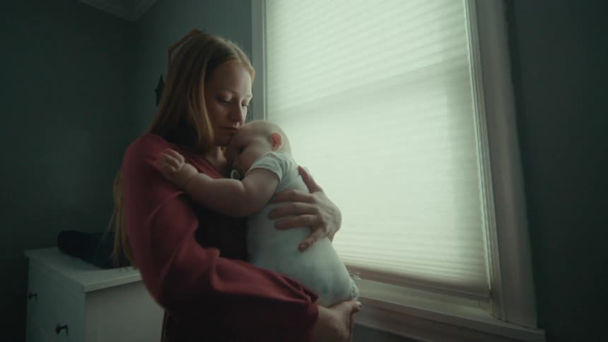 A mother has her small baby on her lap in a white room and is looking at him staring