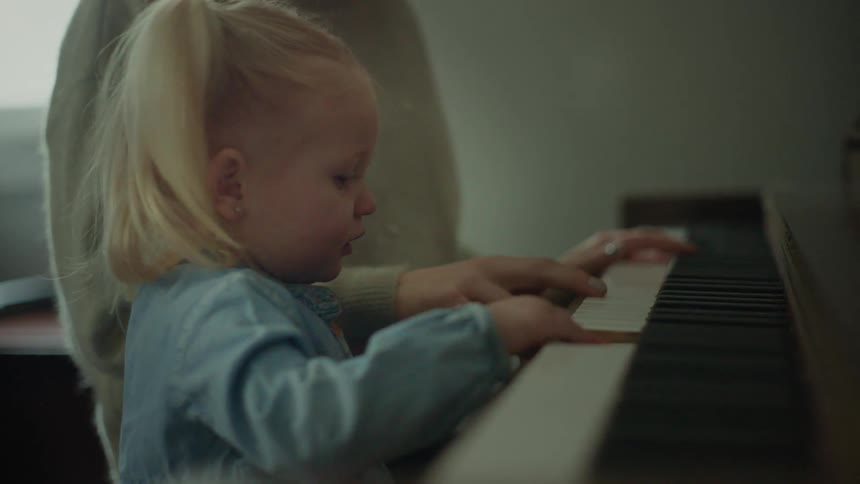 A little blonde girl is playing the piano next to her mother who is looking at her smiling