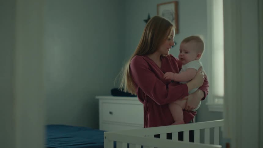 A mother carrying her baby standing next to a crib in a light blue room
