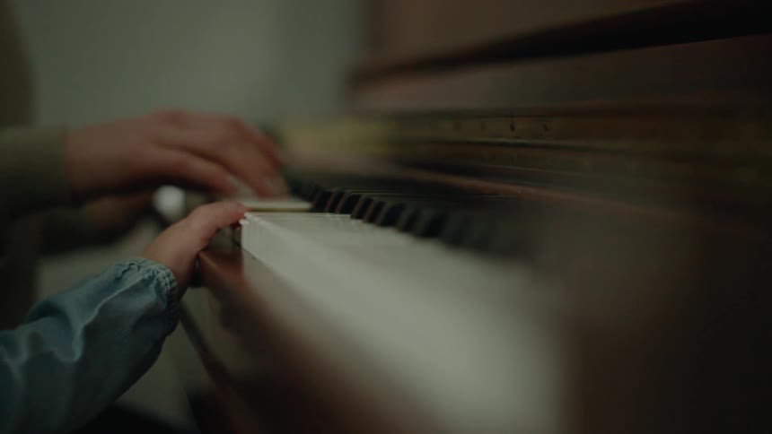 A little blonde girl is playing a piano with the help of an adult next to her