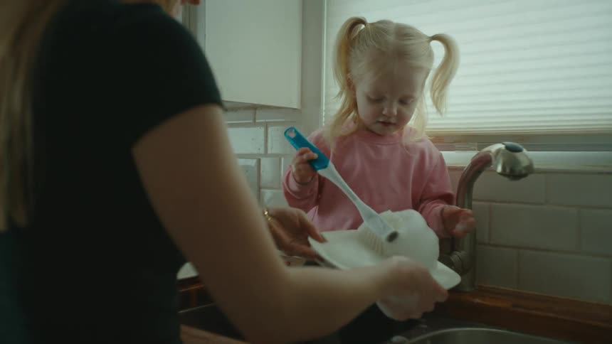 A little blonde girl inside the sink is helping her mother wash the dishes and is catching the suds with her hand