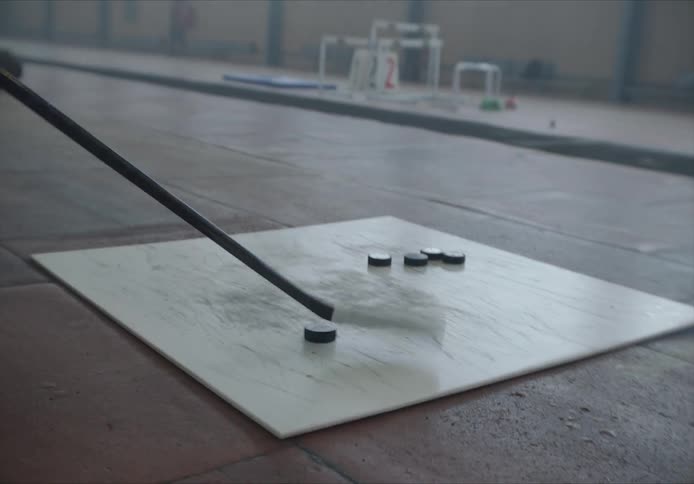 Hockey puck dribbling back and forth with a hockey stick on a synthetic ice rink tile in a building with natural daylight