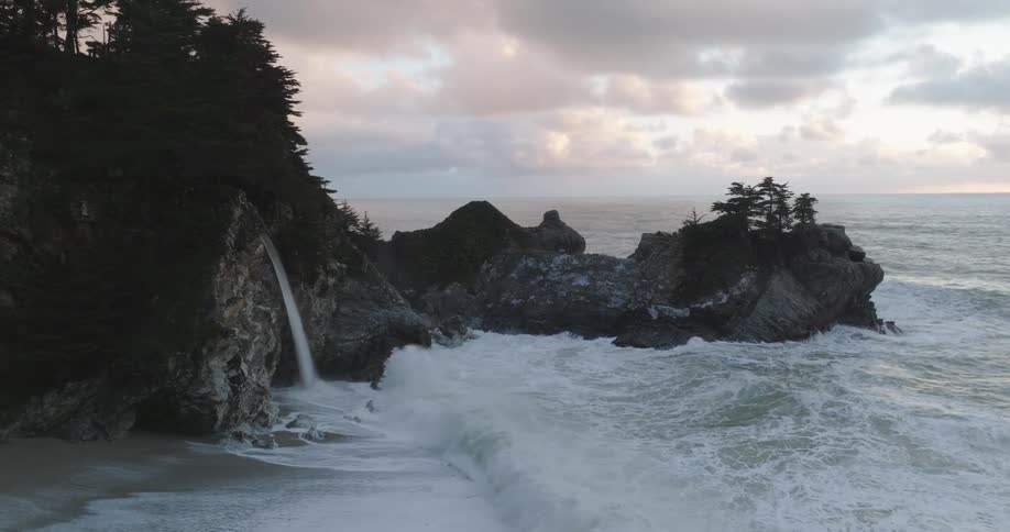 Aerial of waves crashing on mountainous beach shore during sunset