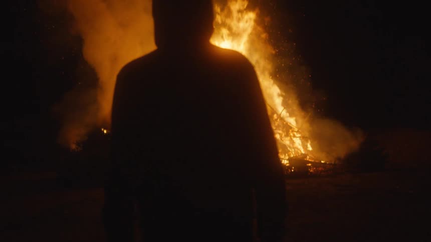 Silhouette of a man wearing a hoodie walking away from a blazing fire