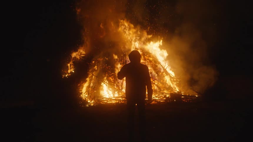 The silhouette of a person can be seen as they stand before a raging fire