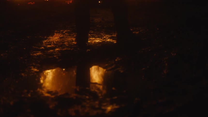 A person stands near a puddle that reflects a raging fire