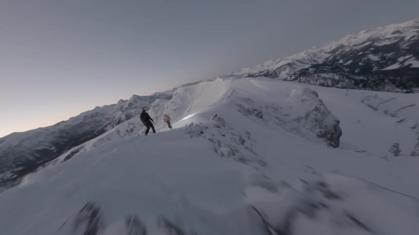An aerial FPV drone shot of two climbers walking on a snowy mountain at dusk
