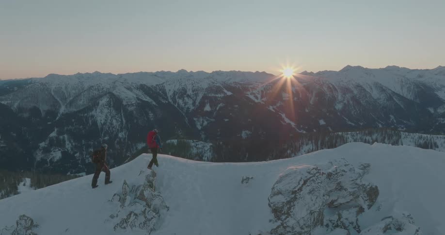 Two hikers on a snowy peak with a sunset view of the mountains