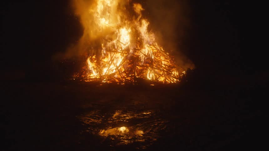 Flames rise as a blazing fire burns in the darkness and reflects on the water puddle below