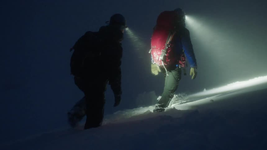 Two men hiking at night in the middle of a snowstorm on a mountain using a led headlamp