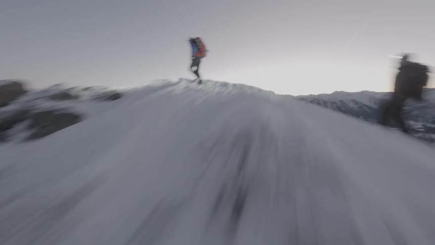 An aerial FPV drone shot zooming by two mountain climbers hiking over a mountain range covered by snow in the morning