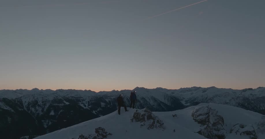 Two hikers on the peak of a mountain covered by snow in a dark morning sky
