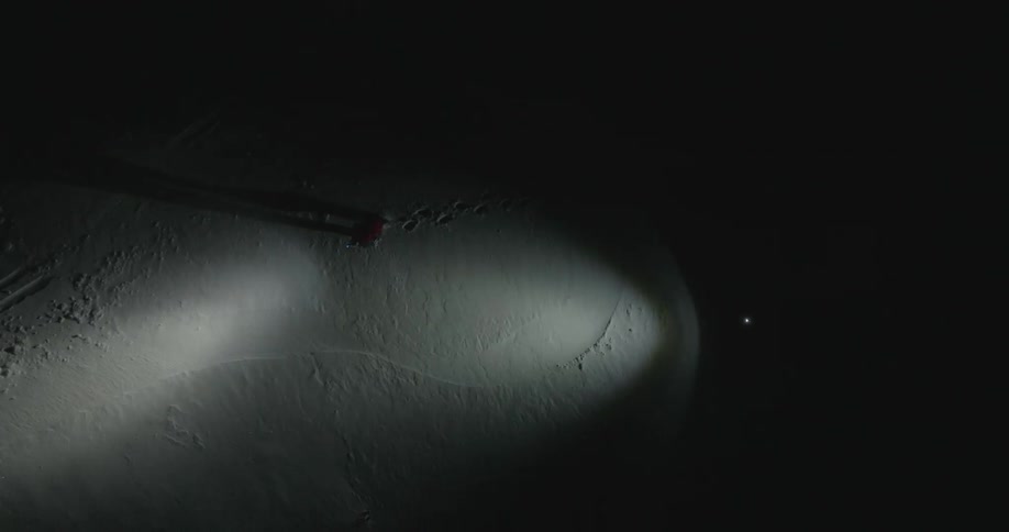 Two people walking on a dark snowy landscape illuminated by flashlights at night