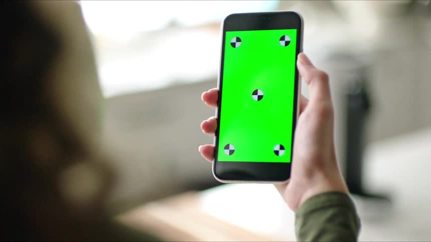 A woman holds a cell phone with a green screen as she drinks from a cup