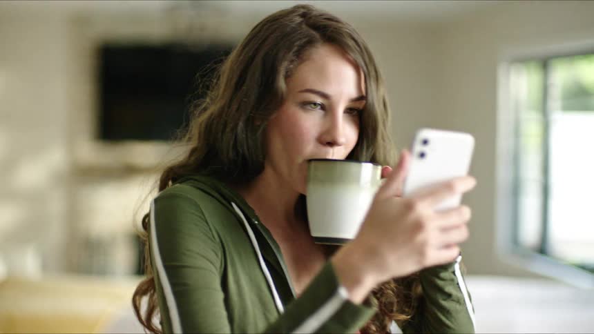 Portrait of a young woman holding a cup of coffee and her cell phone chatting while walking around the room