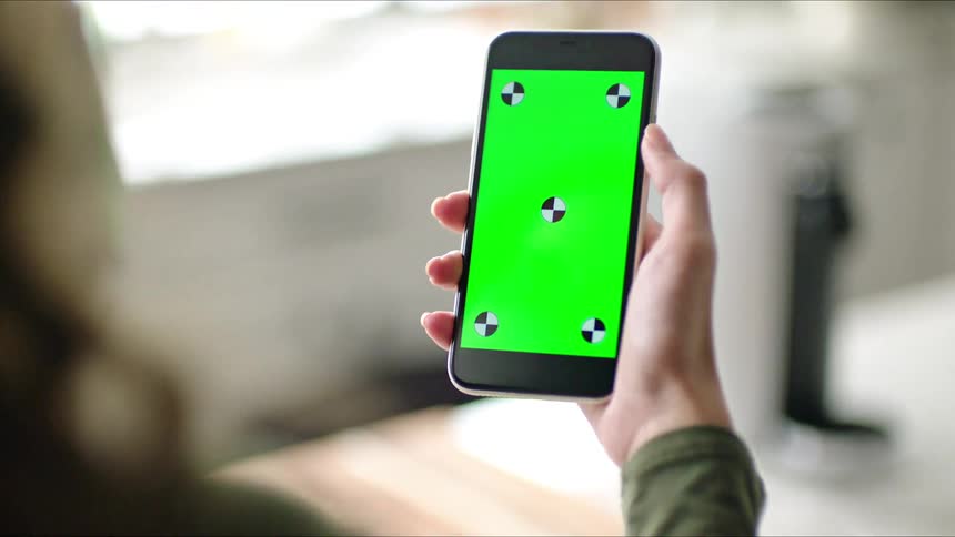 A woman holds a cell phone with a green screen as she drinks from a cup