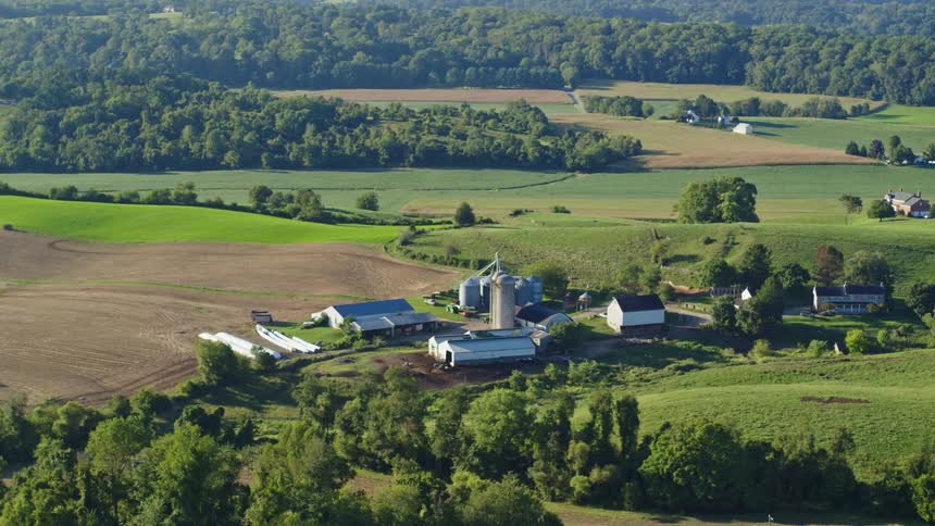A farm with a barn in a silo in a region with large fields of grass and several trees back