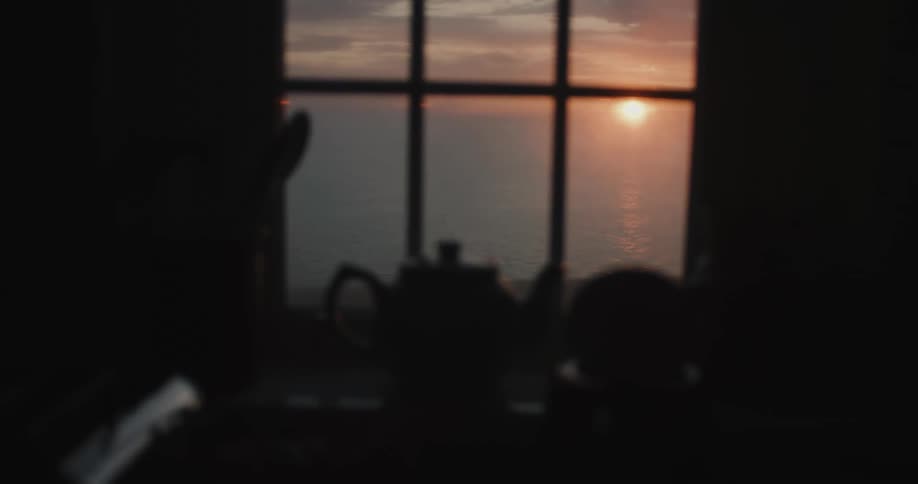 Teapot near window stands silhouetted against ocean and the sun setting on horizon