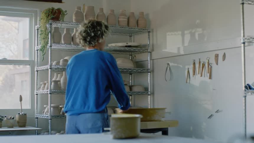 A blonde person in a blue shirt takes an object from the table and places it on another surface