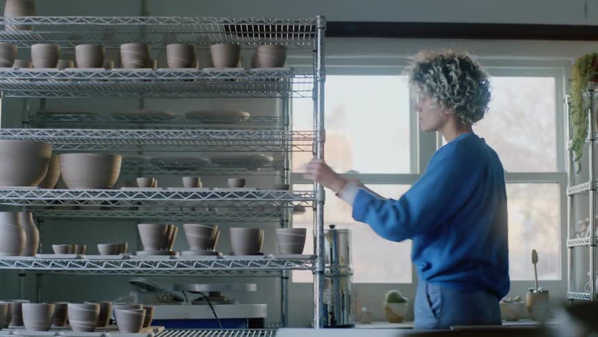 Female potter moves clay pots from shelves and onto the table