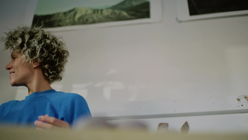 A blonde woman with short hair and wearing a blue shirt is holding a pot of clay and looks back and then lets out a smile