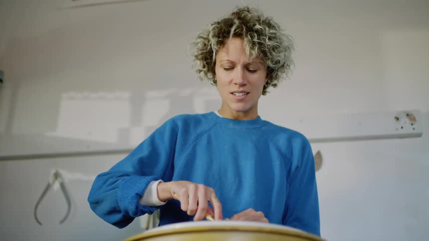 A woman is speaking while using tools to shape a piece of clay on a pottery wheel