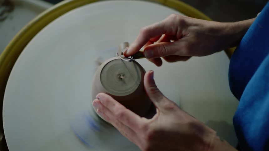 A persons hands that are molding a small clay pot with a tool and a turntable