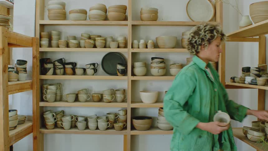 A person has their back turned wearing a green jacket and is organizing clay pots on a shelf
