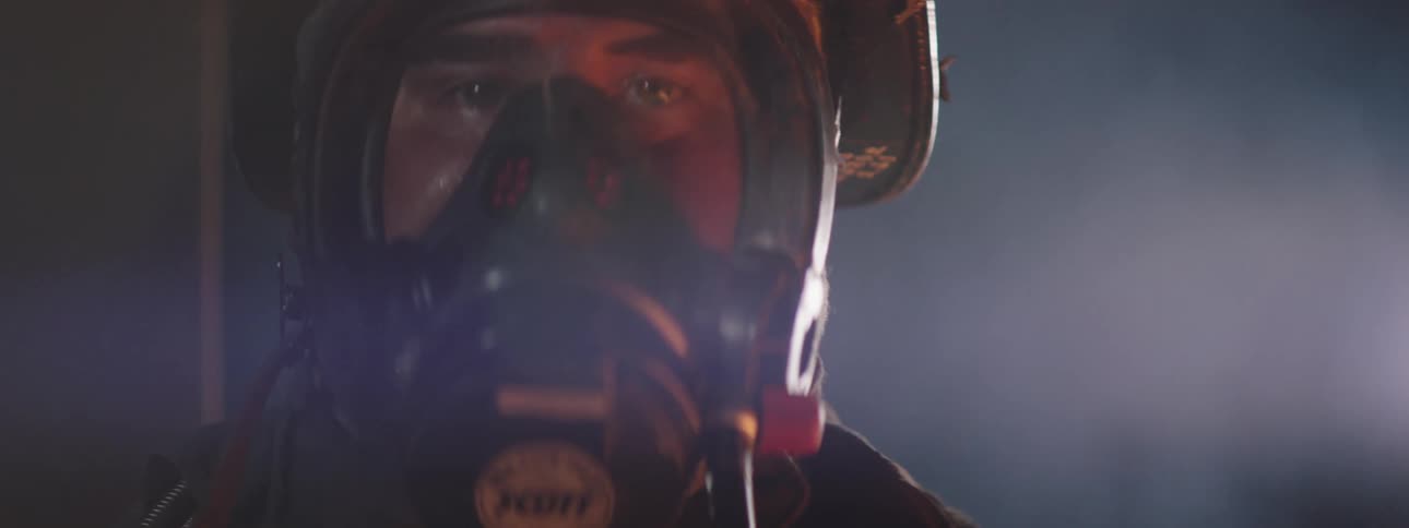 A firefighter wearing a mask looks directly into the camera as smoke rises behind him