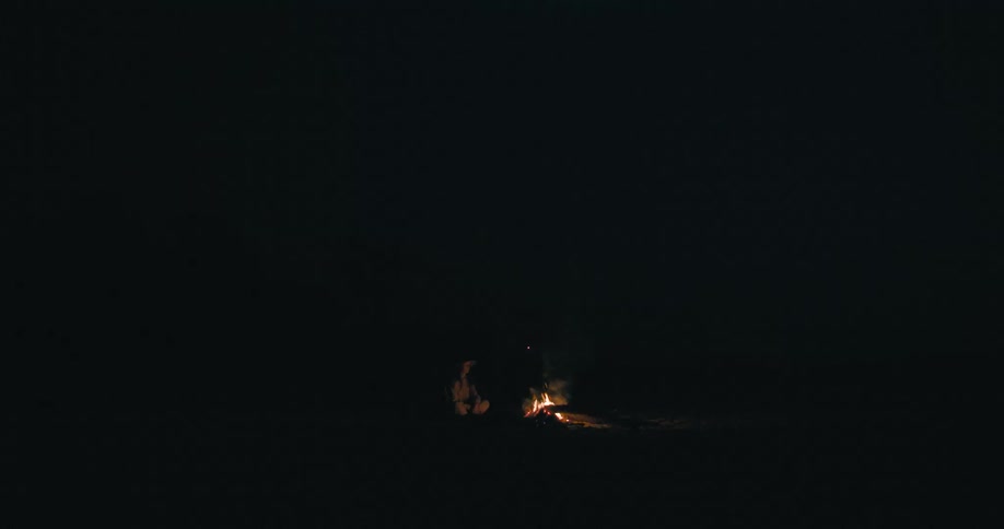 A person sits in the darkness next to a burning campfire