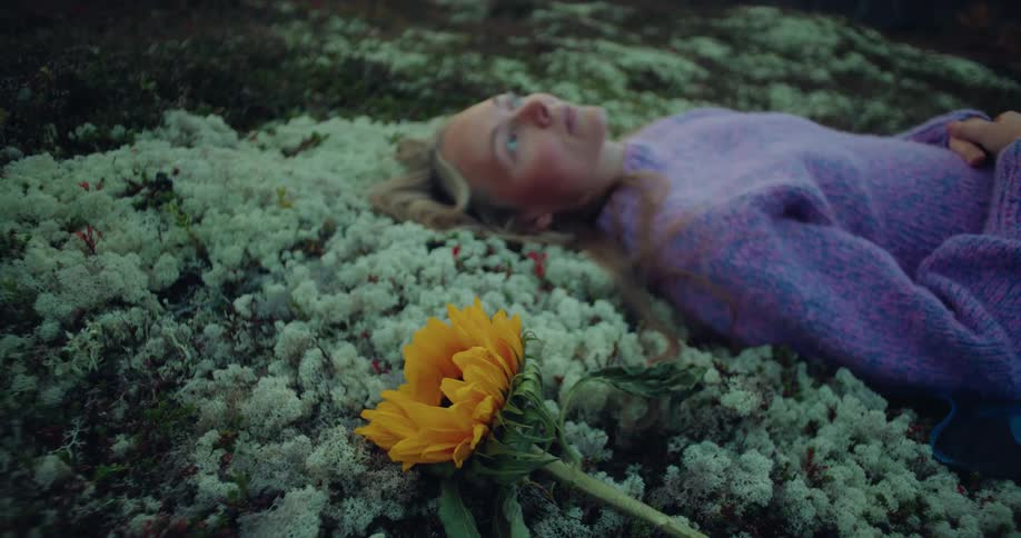A blonde woman in purple lying on a mossy ground with a yellow flower