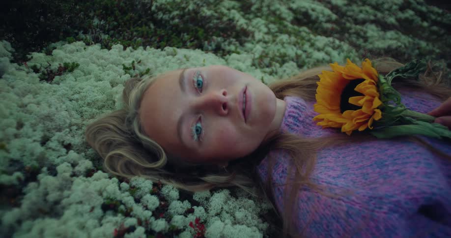 A woman lays on her back on the ground while holding a sunflower to her chest