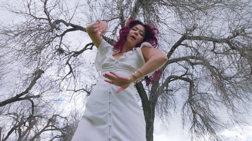 Woman with red hair and white dress dances under bare tree on a cloudy day