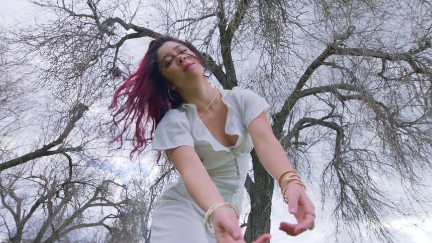 Woman in a white dress shakes her long colorful hair in front of a barren tree